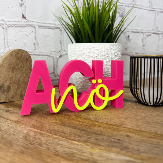 3D-Schriftzug „Ach nö“ in Rosa-Gelb als humorvolle Deko auf Holztisch