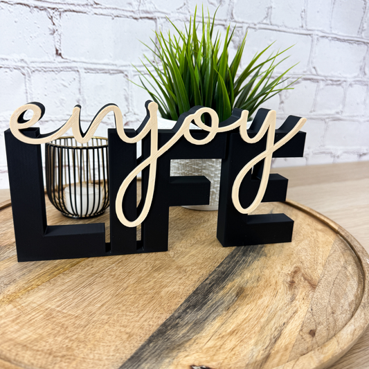 3D Schriftzug „LIFE“ in Schwarz mit beigefarbenem Schriftzug „enjoy“, frontale Ansicht auf Holzuntergrund mit Pflanze im Hintergrund