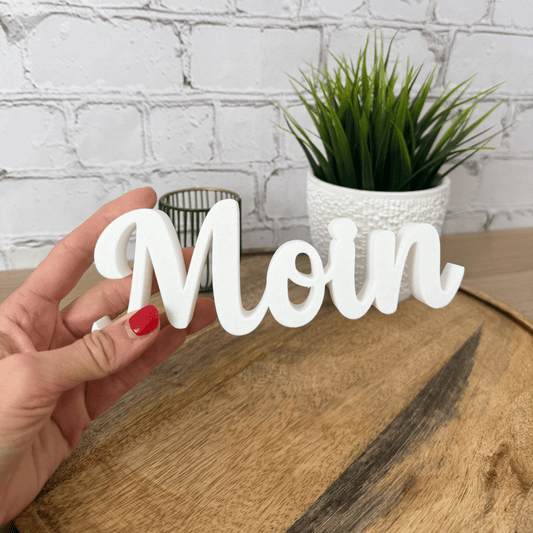 3D Schriftzug „Moin“ in Weiß, aus PLA gefertigt, in der Hand gehalten auf Holzfläche vor heller Ziegelwand