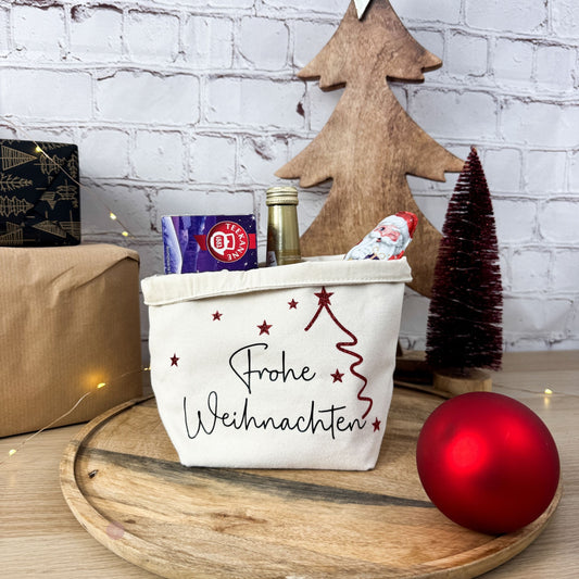 Ansicht des Geschenkkorbs „Frohe Weihnachten“ auf einem Holzbrett, gefüllt mit Tee und Sekt. Im Hintergrund ein roter Tannenbaum und ein roter Weihnachtsstern.