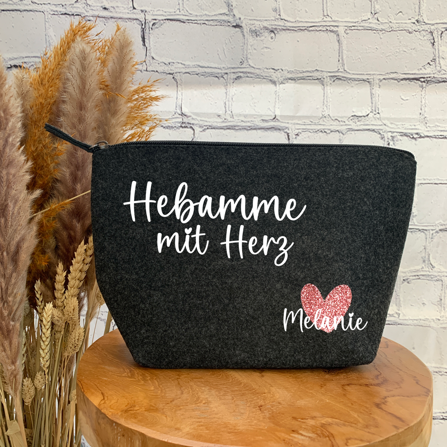 Filz-Kulturtasche in Anthrazit mit dem Schriftzug „Hebamme mit Herz“ und einem glitzernden pinken Herz mit dem Namen „Melanie“.