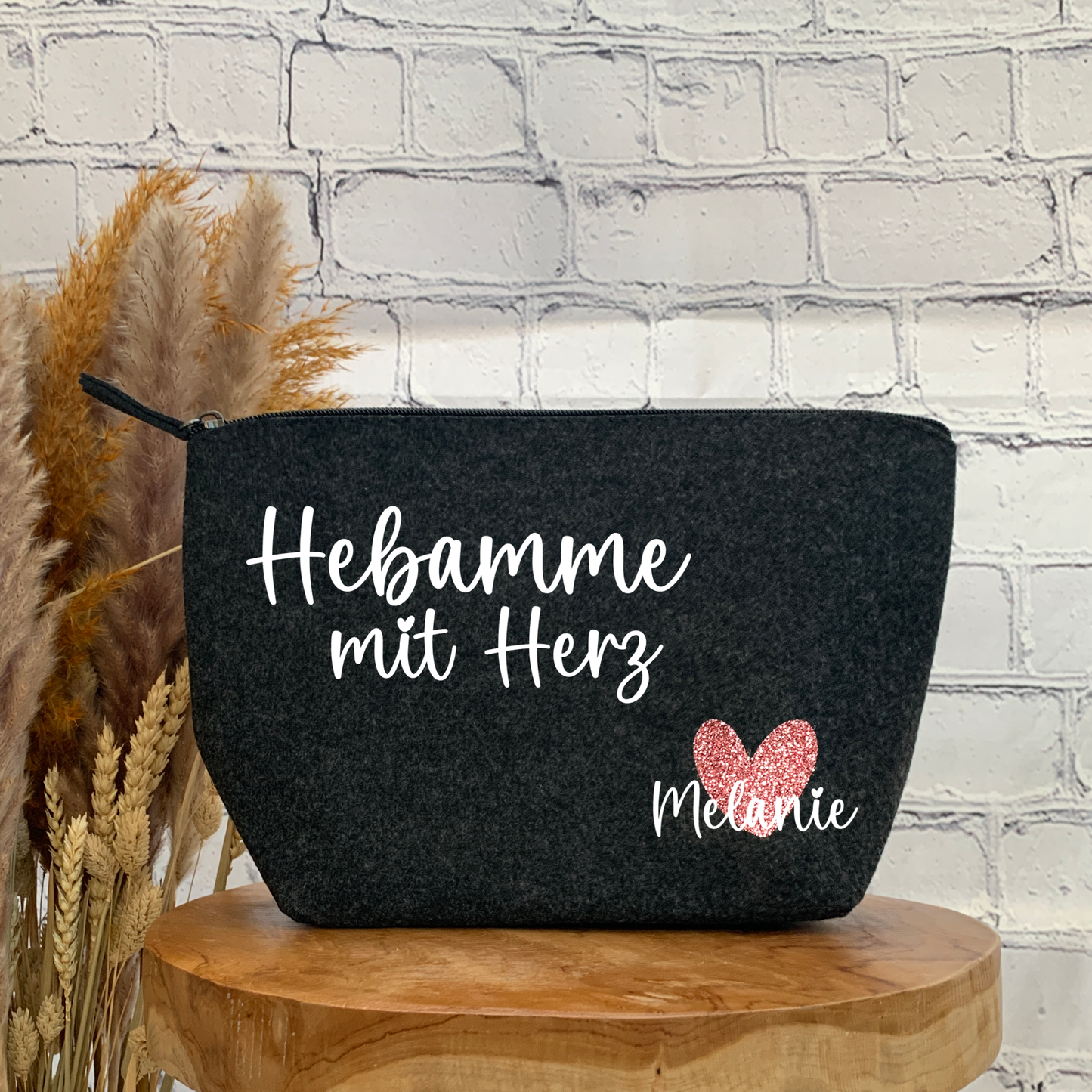 Nahaufnahme einer anthrazitfarbenen Filz-Kosmetiktasche mit dem weißen Schriftzug „Hebamme mit Herz“ und einem glitzernden pinken Herz, personalisiert mit dem Namen „Melanie“.