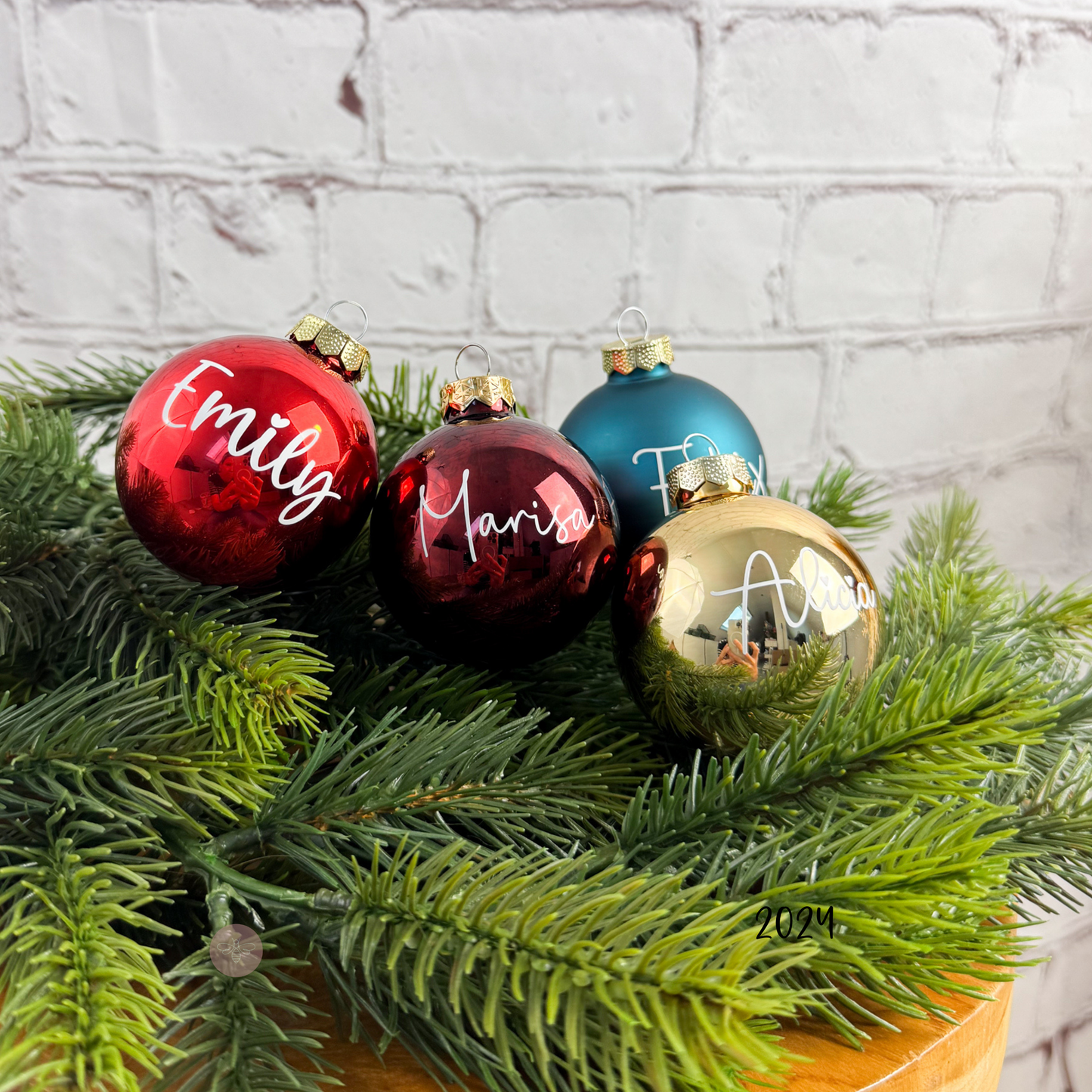 Vier personalisierte Weihnachtskugeln mit Namen in verschiedenen Farben und Schriften auf Tannenzweigen – das perfekte Geschenk zur Weihnachtszeit.