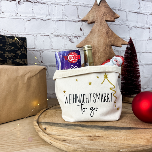 Ansicht des Geschenkkorbs "Weihnachtsmarkt to go" auf einem Holzbrett, gefüllt mit Tee und Sekt. Im Hintergrund ist ein Tannenbaum zu sehen.