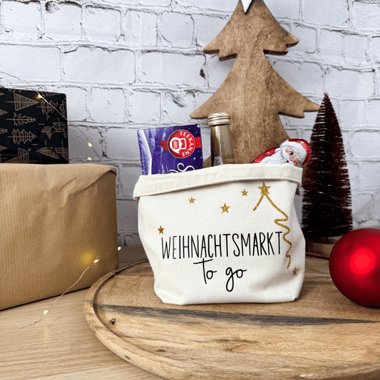Ansicht des Geschenkkorbs "Weihnachtsmarkt to go" auf einem Holzbrett, gefüllt mit Tee und Sekt. Im Hintergrund ist ein Tannenbaum zu sehen.