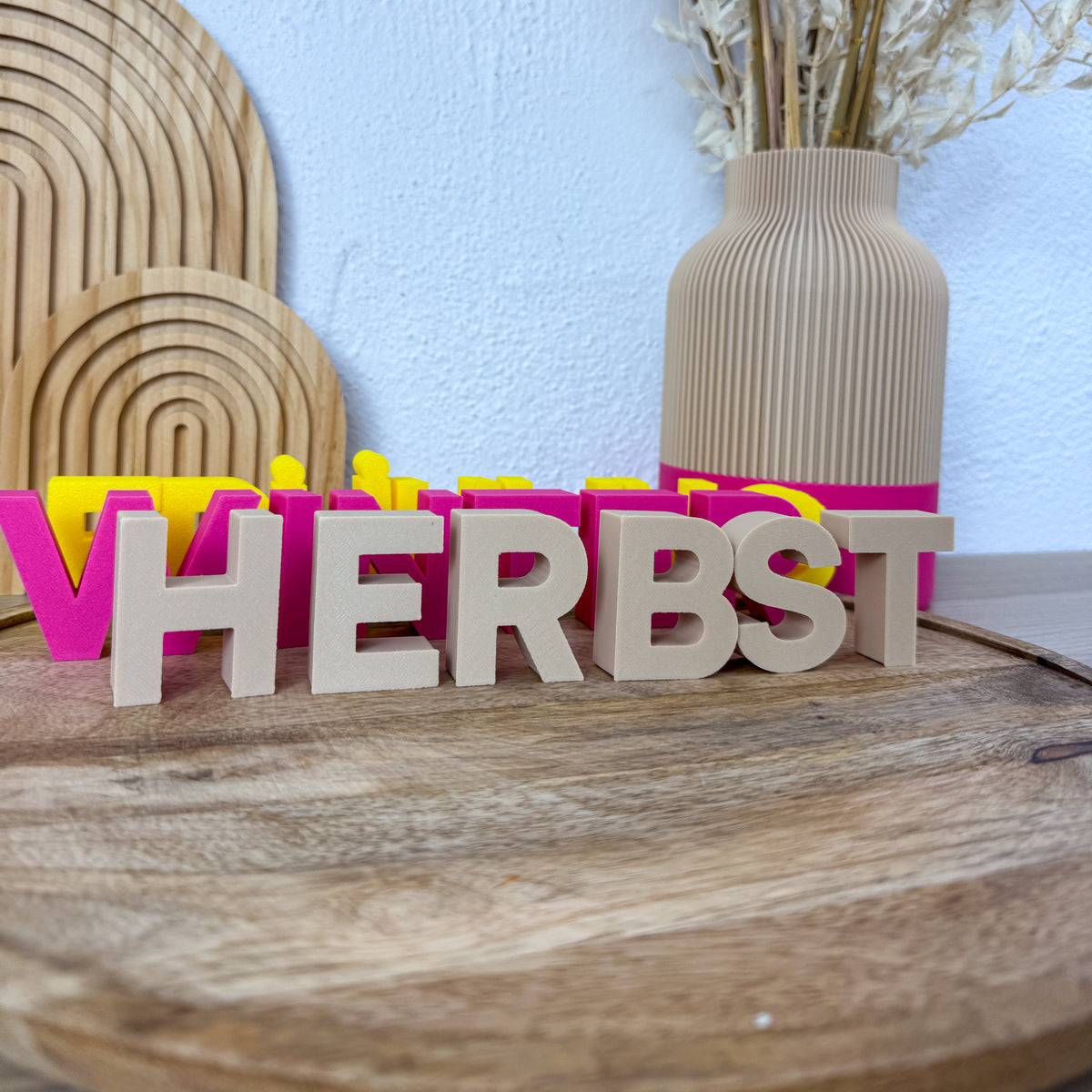 Deko Buchstaben HERBST als 3D-Druck in Beige, Pink und Gelb nebeneinander auf einem runden Holzbrett