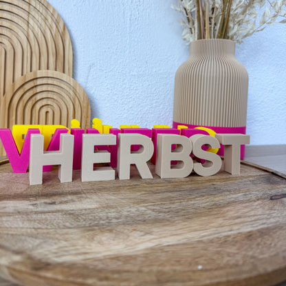Deko Buchstaben HERBST als 3D-Druck in Beige, Pink und Gelb nebeneinander auf einem runden Holzbrett