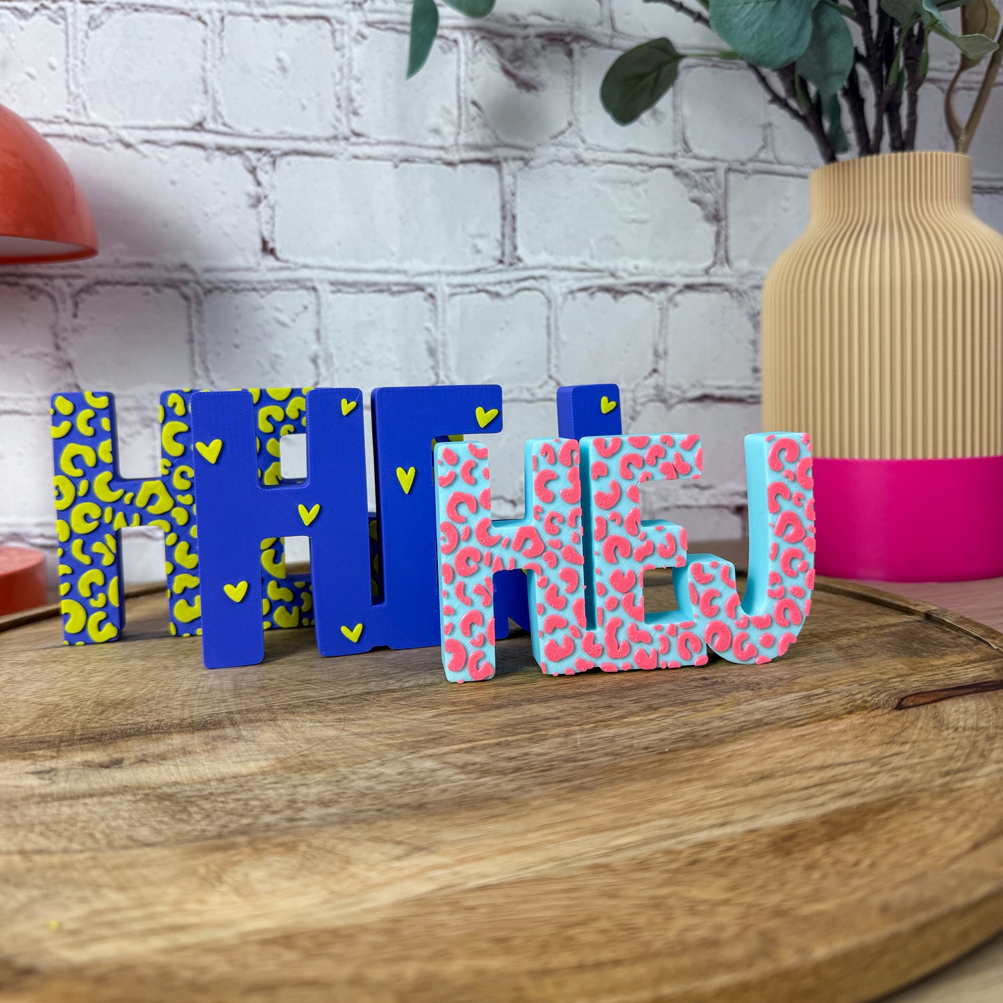 Vier 3D-Schriftzüge HEJ nebeneinander in Blau-Gelb und Türkis-Pink mit Leo- und Herzmuster