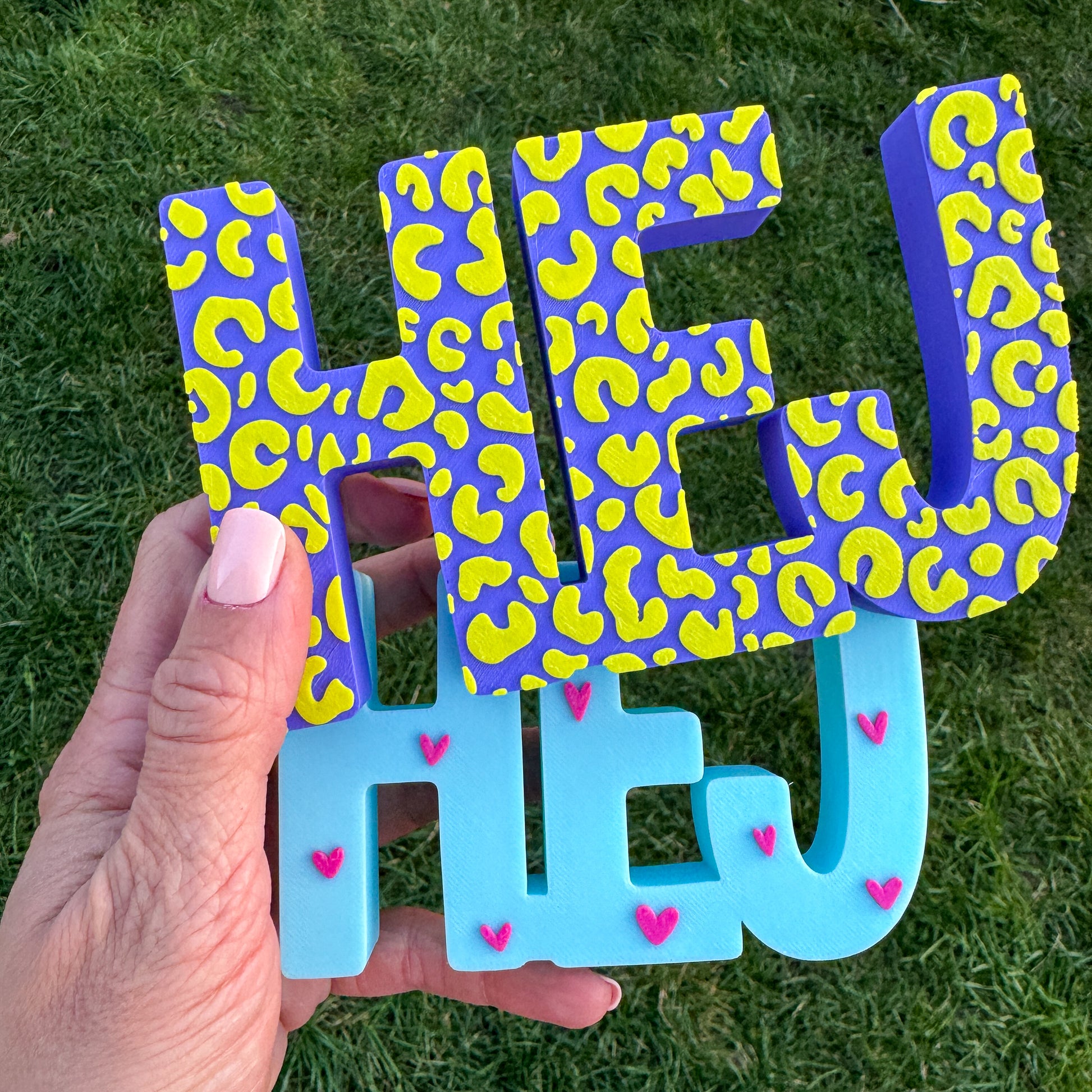 3D-Schriftzug HEJ in Blau-Gelb-Leo und Türkis-Pink-Herzen, im Freien gehalten