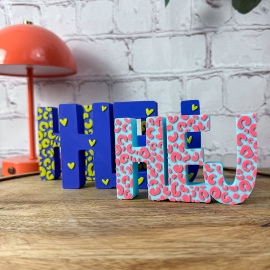 Drei 3D-Schriftzüge HEJ in Blau-Gelb und Türkis-Pink mit Leo-Muster auf Holztablett