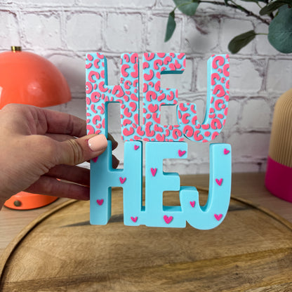 3D-Schriftzug HEJ in Türkis, oben pinkes Leo-Muster unten pinke Herzchen, in Hand