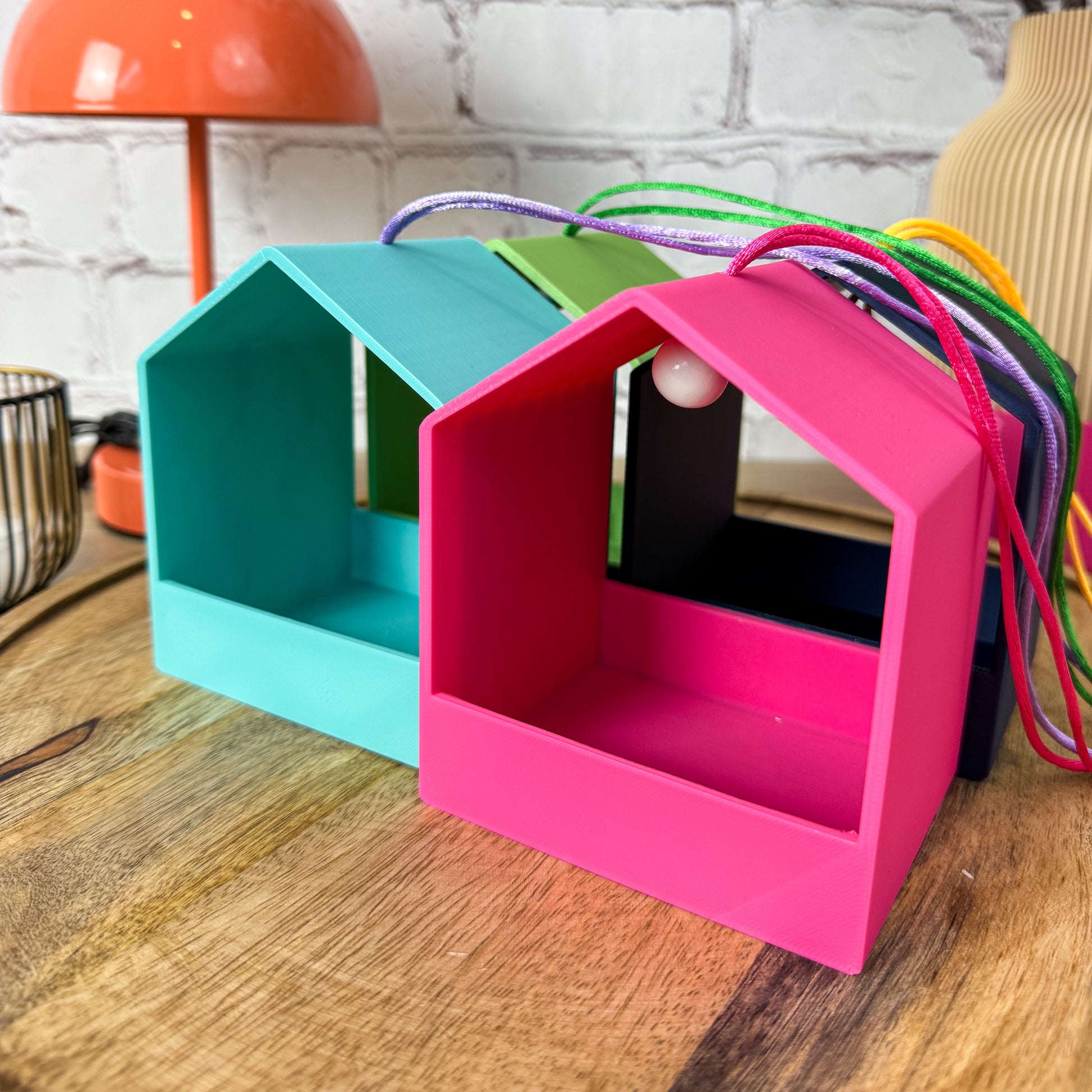 Nahaufnahme 3D-gedruckter Vogelfutterstationen in Türkis und Pink mit bunten Aufhängebändern