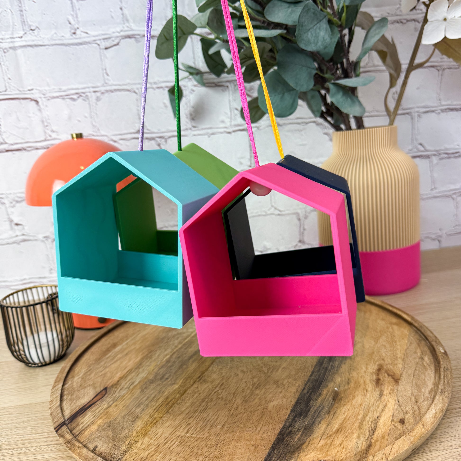 Hängende Vogelfutterstationen aus PLA in Türkis und Pink mit farbigen Bändern in der Luft