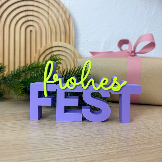 3D Schriftzug „frohes Fest” in Lila und Neongelb vor einem Geschenk mit rosa Schleife