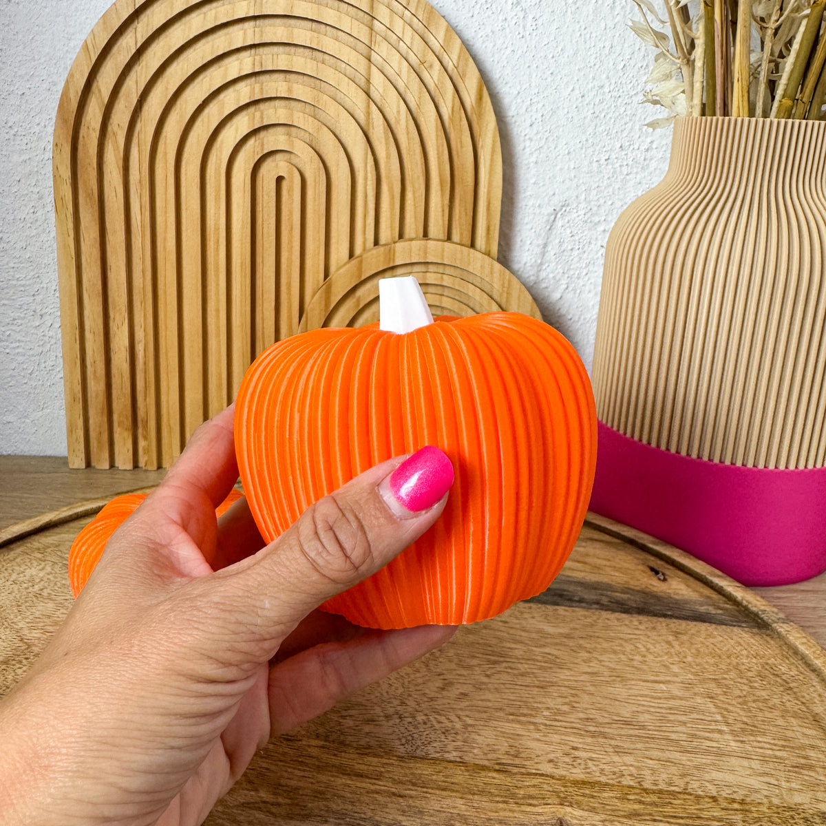Großer 3D Kürbis Skandi in Orange in einer Hand gehalten, Größenvergleich vor Holzbögen und Vase
