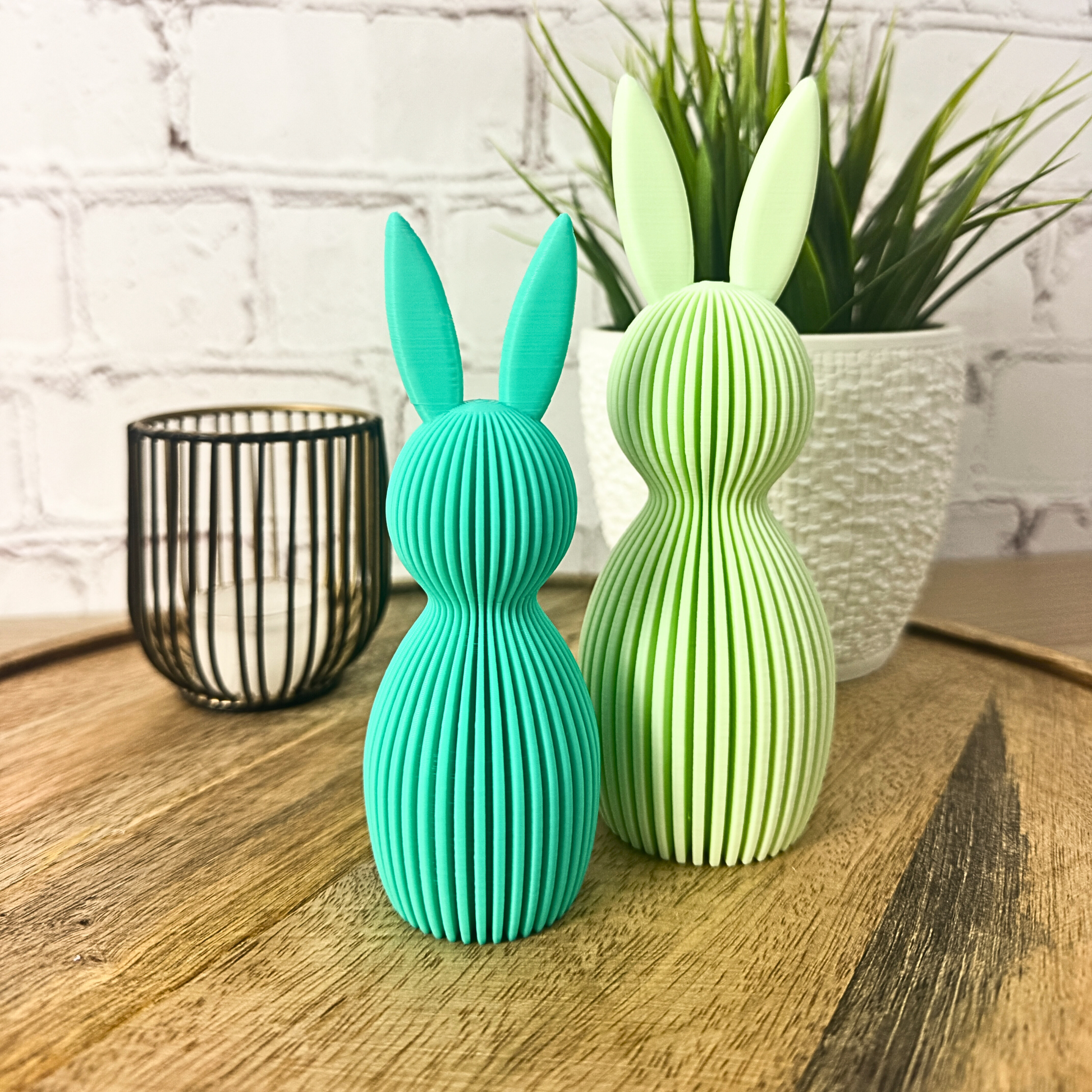 Zwei geriffelte Osterhasen aus PLA in Türkis und Hellgrün, stehend auf Holzuntergrund vor Ziegelwand