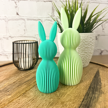 Zwei geriffelte Osterhasen aus PLA in Türkis und Hellgrün, stehend auf Holzuntergrund vor Ziegelwand