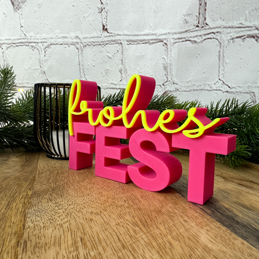 3D Schriftzug 'frohes Fest' in Pink mit gelber Schrift, dekorativ auf Holztisch mit Tannenzweigen arrangiert