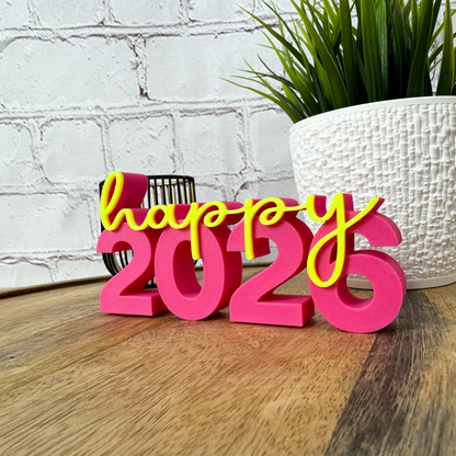 3D Schriftzug „happy 2026“ aus PLA in Pink und Gelb als Silvester-Deko, dekorativ auf Holztisch vor weißer Wand