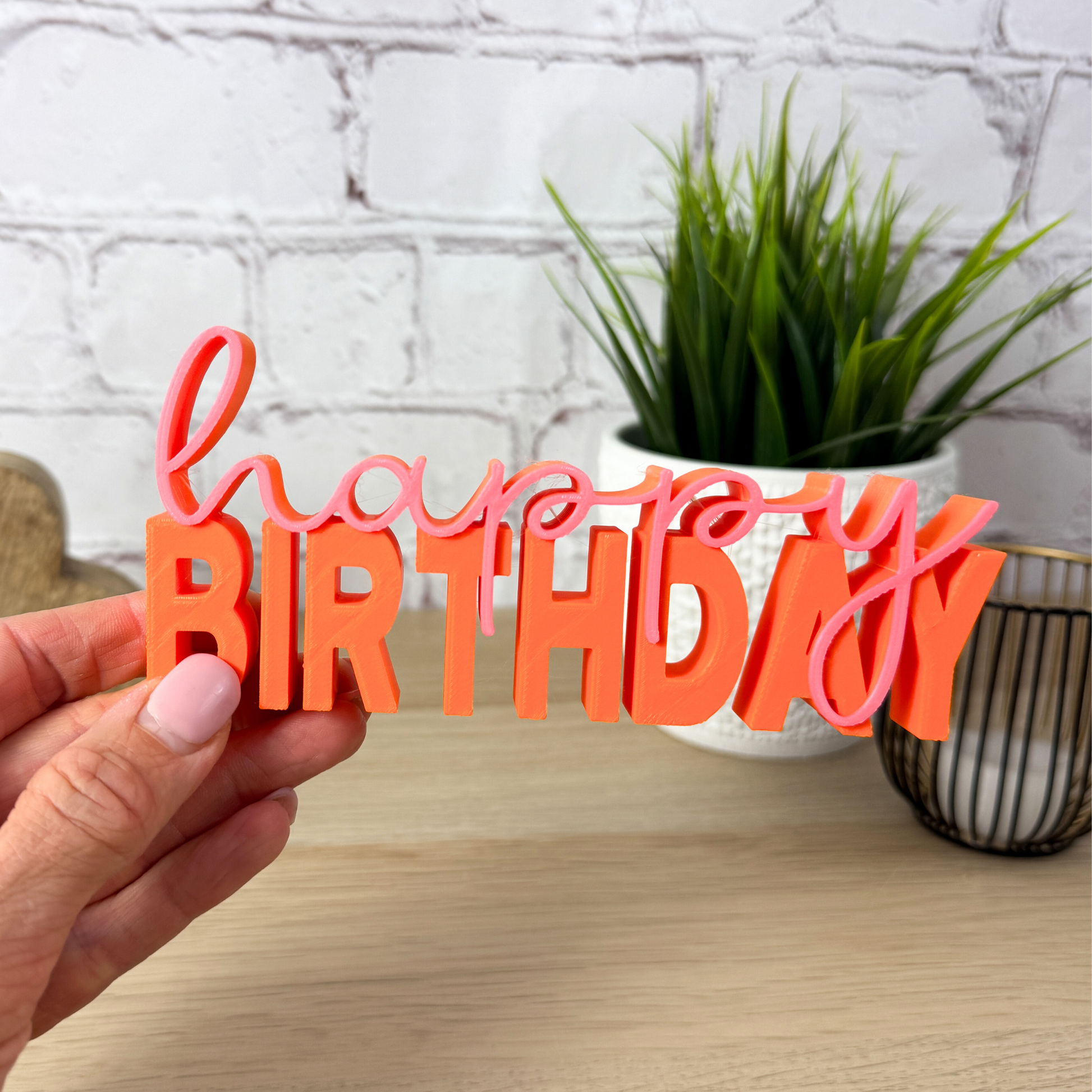Orange-pinker 3D-Schriftzug „happy birthday“ aus PLA, liebevoll dekoriert auf Holztisch vor Pflanze