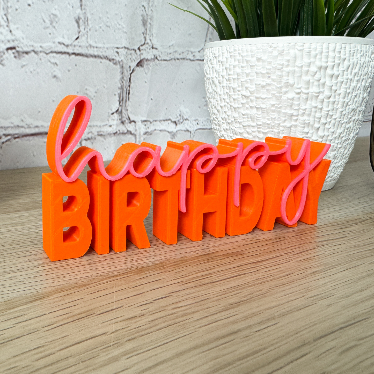 Orange-pinker 3D-Schriftzug „happy birthday“ aus PLA+, dekorativ vor weißem Blumentopf auf Holztisch