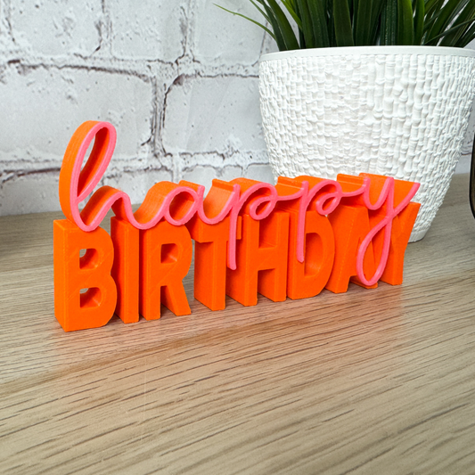 Orange-pinker 3D-Schriftzug „happy birthday“ aus PLA+, dekorativ vor weißem Blumentopf auf Holztisch