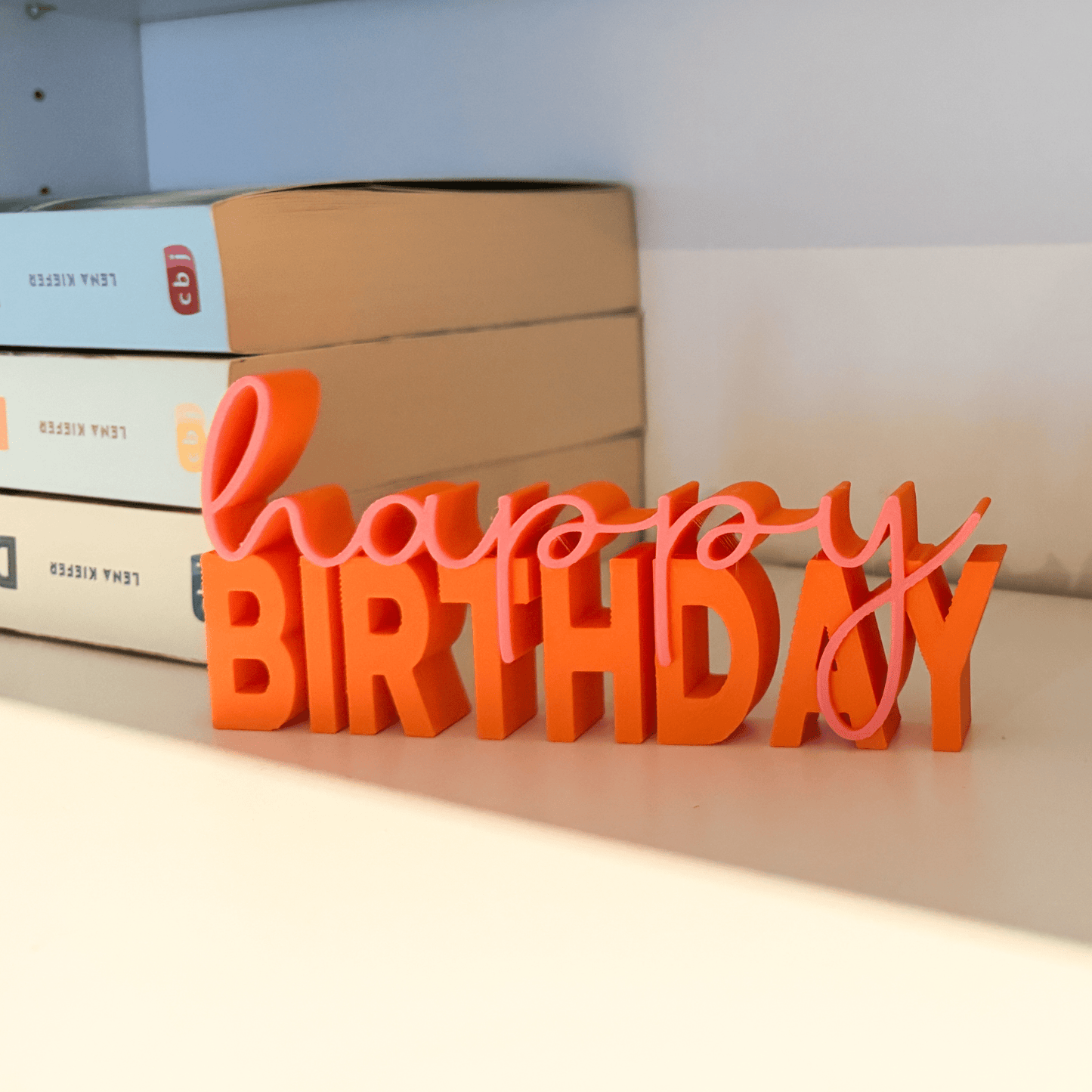 3D-Schriftzug „happy birthday“ in Orange und Pink als Regal-Deko neben Büchern, ideal für Geburtstage