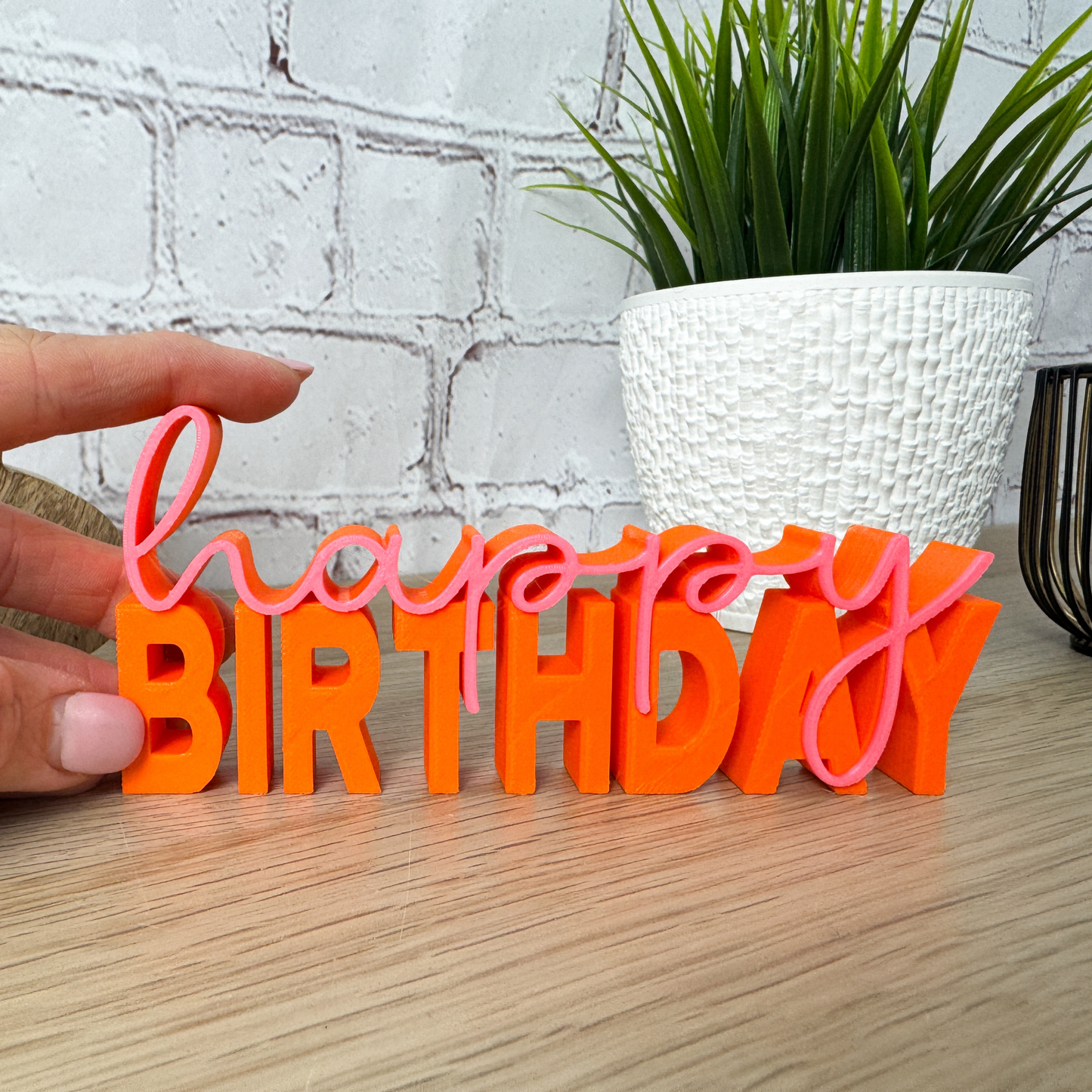 Hand hält orange-pinken 3D-Schriftzug „happy birthday“ aus PLA, dekorativ vor Pflanze und Dekoobjekten