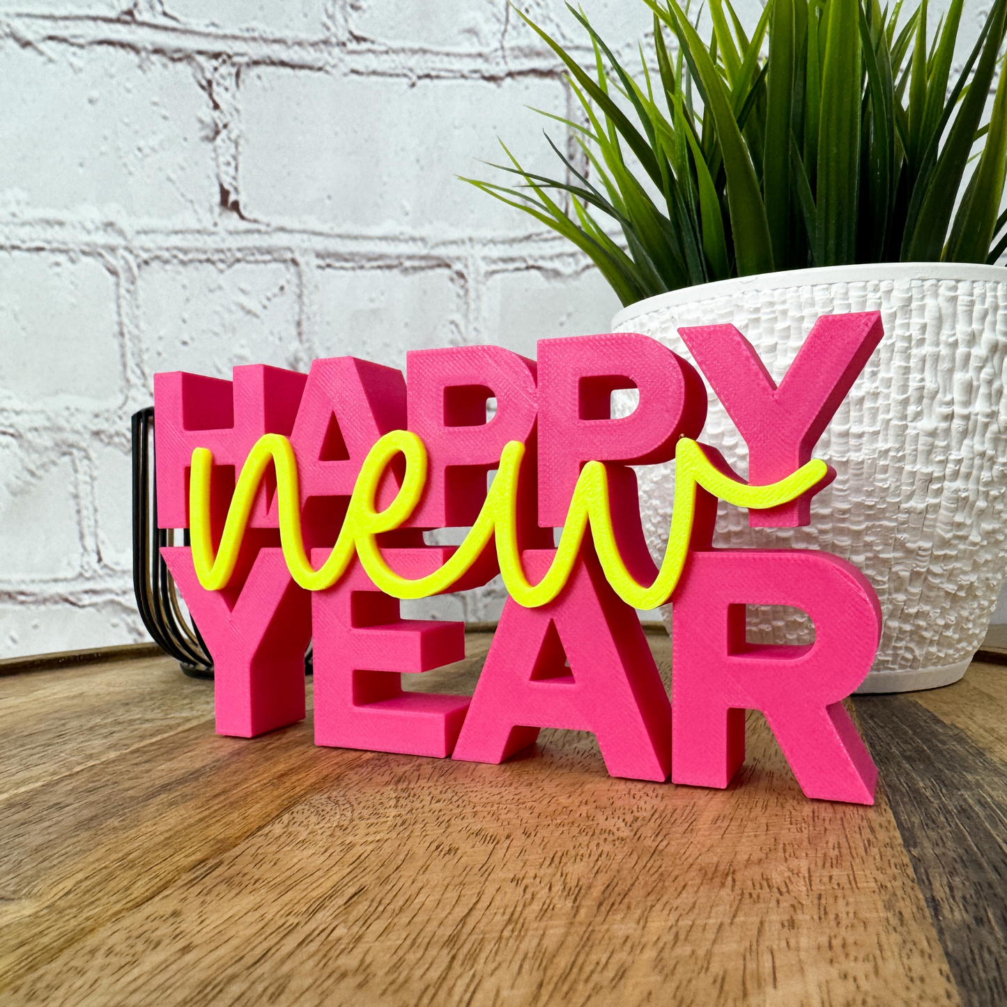 PLA 3D Schriftzug „happy new year“ in leuchtendem Pink und Gelb, dekorativ vor Pflanze und Backsteinwand