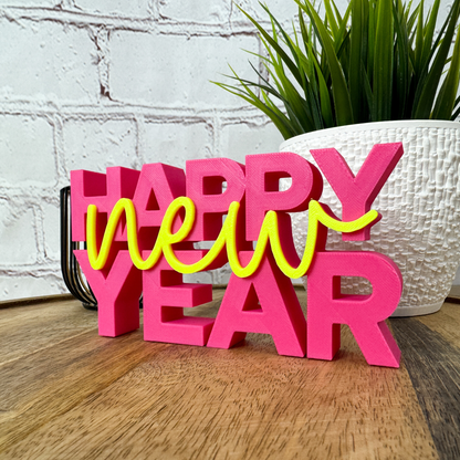 PLA 3D Schriftzug „happy new year“ in leuchtendem Pink und Gelb, dekorativ vor Pflanze und Backsteinwand