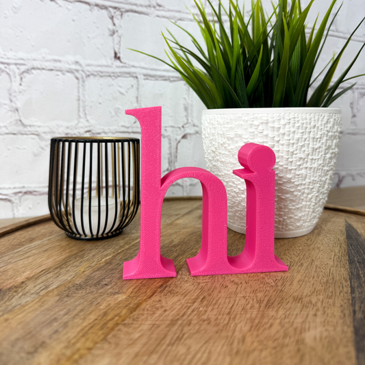 Pinker 3D Schriftzug „hi“ aus PLA als dekorativer Deko Schriftzug auf Holztisch neben Pflanze und Kerze