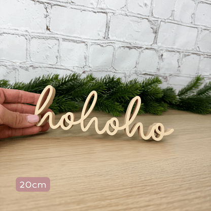 hohoho 3D-Schriftzug in Beige, 20 cm breit, in der Hand gehalten vor Tannengirlande und Backsteinwand