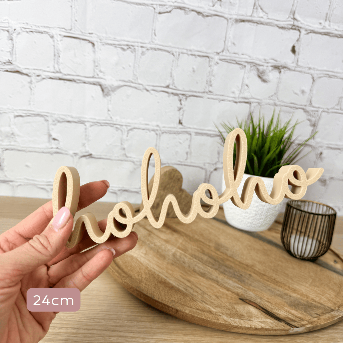 hohoho Schriftzug in Beige, 24 cm breit, in einer Hand vor Holzbrett mit Pflanze und Teelicht