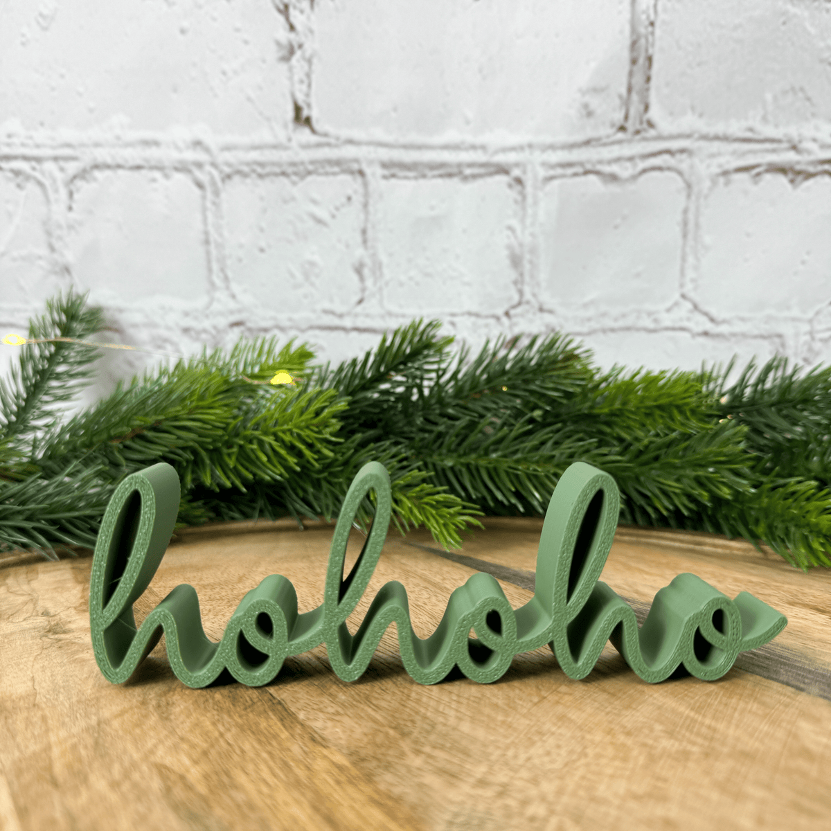 hohoho 3D-Schriftzug in Dunkelgrün auf einem Holzbrett vor Tannengirlande und weißer Backsteinwand