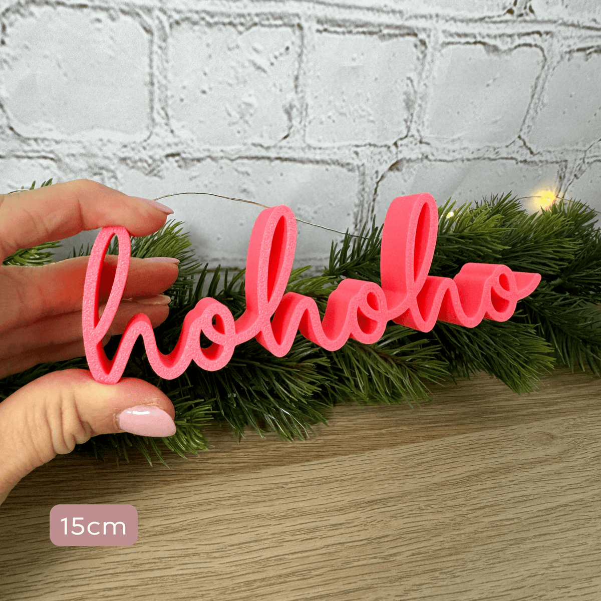 hohoho Schriftzug in Rosa, 15 cm breit, in einer Hand als Größenvergleich vor Tannengrün