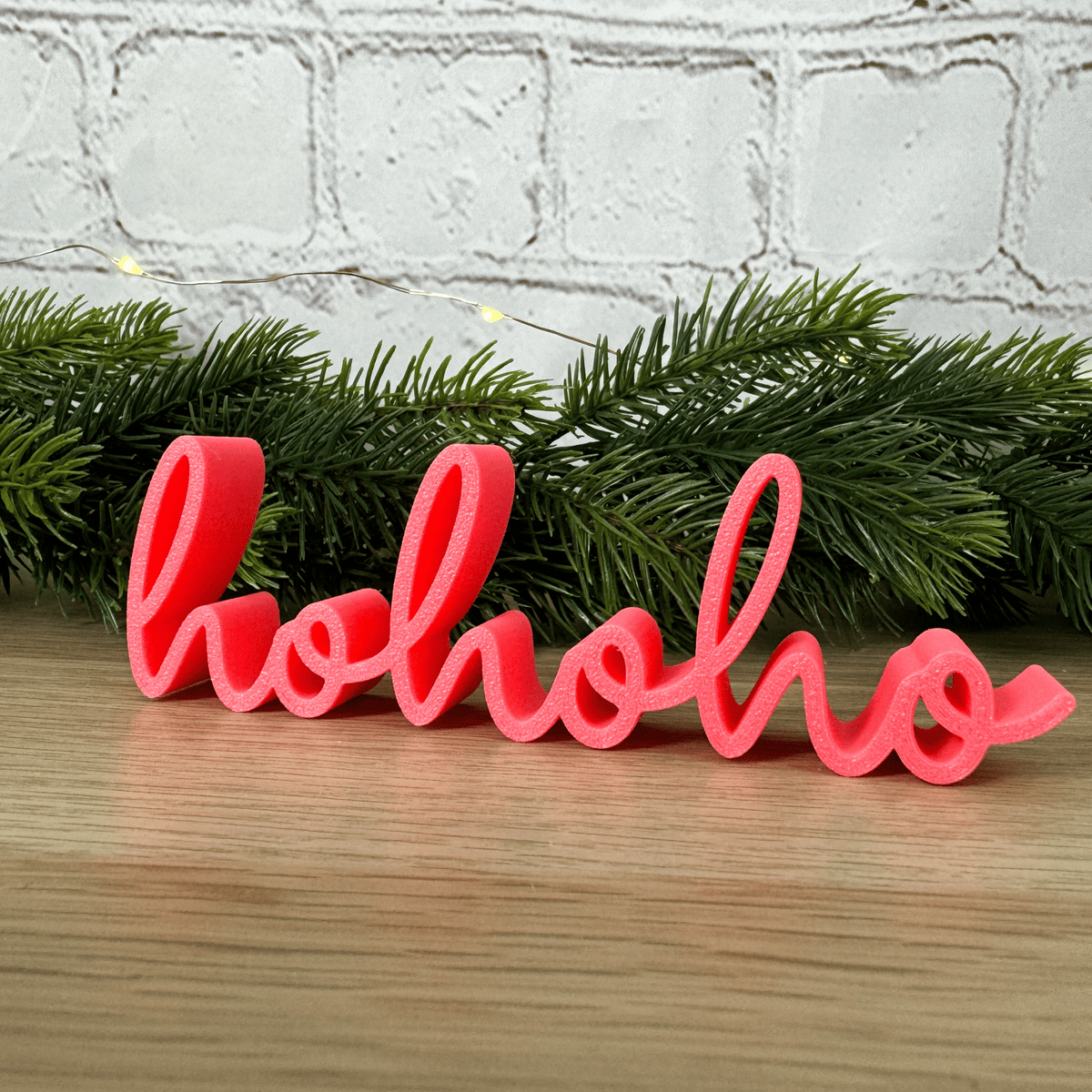 hohoho Schriftzug in Rosa vor Tannenzweigen und Lichterkette, aufgestellt auf einer Holzfläche