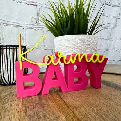 3D Schriftzug „Karma Baby“ aus PLA in Pink mit gelber Schrift, dekorativer Spruch auf Holztisch
