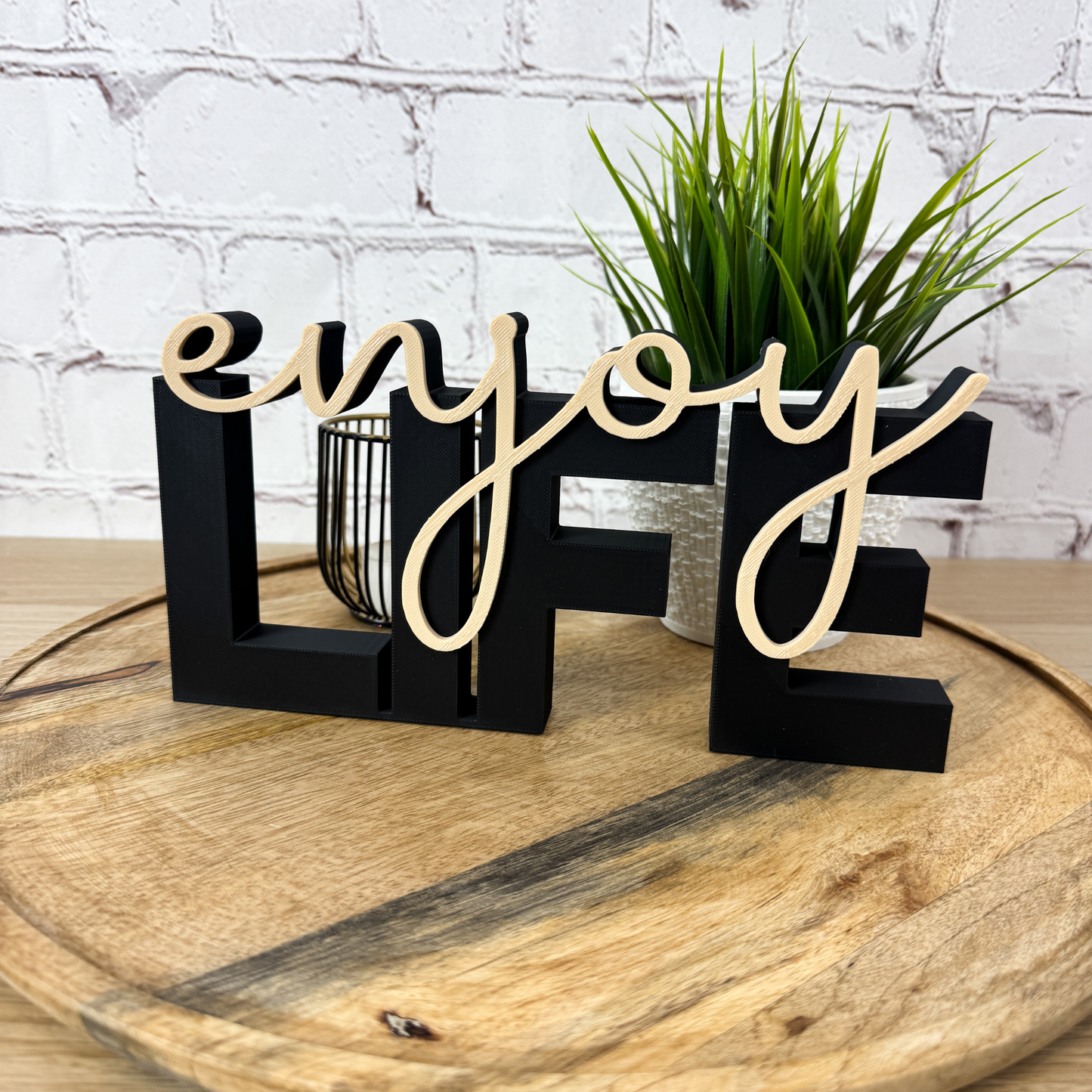 Schwarzer 3D Schriftzug „LIFE“ aus PLA mit aufgesetztem Wort „enjoy“ in Beige, stehend auf rundem Holztablett vor Ziegelwand