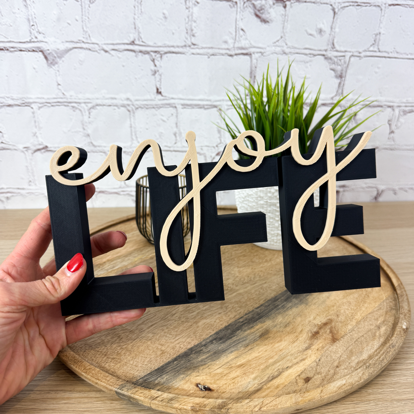 Schwarzer 3D Schriftzug „LIFE“ mit beigefarbenem Wort „enjoy“, gehalten in der Hand auf Holztablett vor weißer Wand