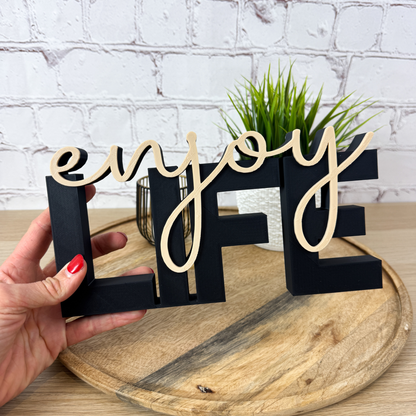 Schwarzer 3D Schriftzug „LIFE“ mit beigefarbenem Wort „enjoy“, gehalten in der Hand auf Holztablett vor weißer Wand