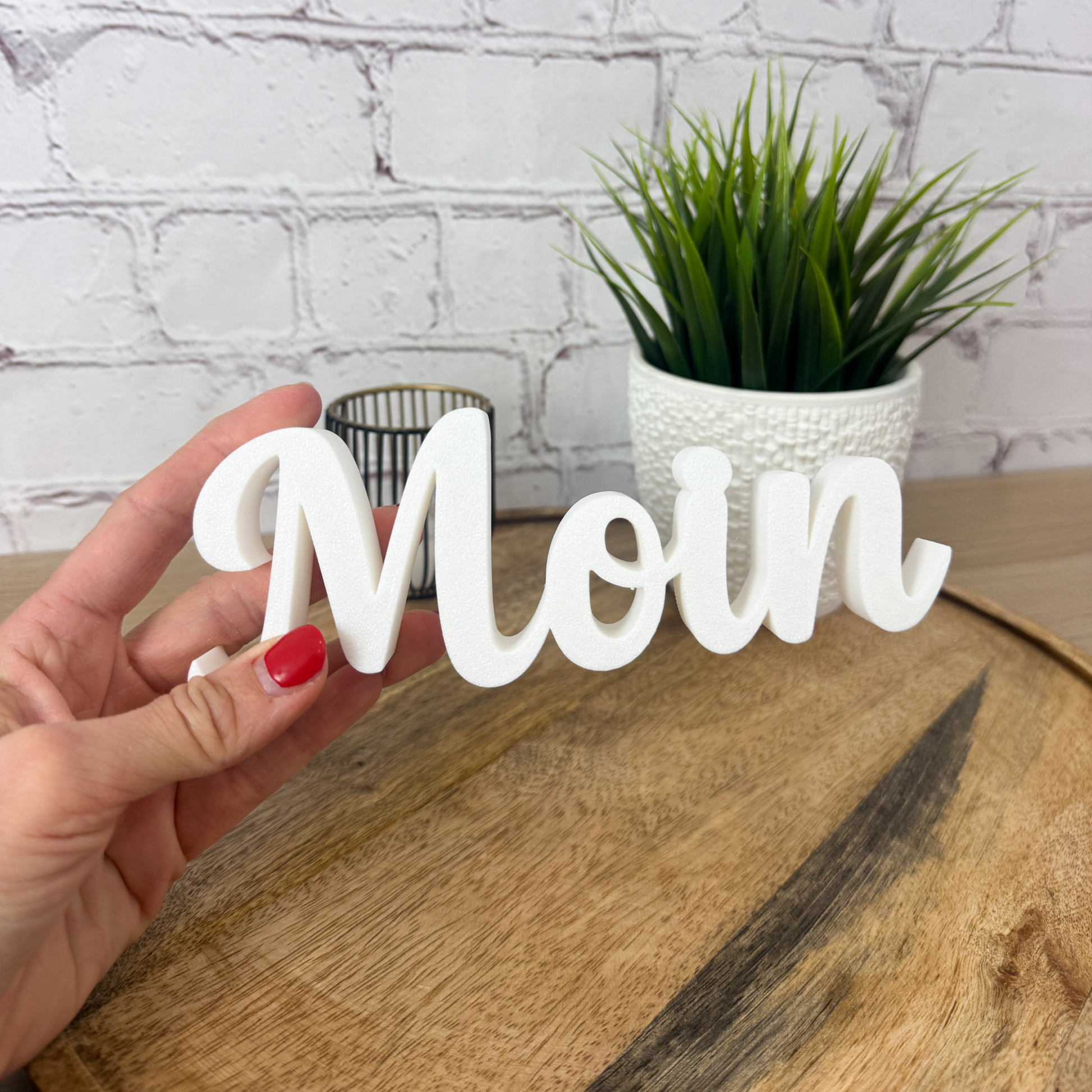 3D Schriftzug „Moin“ in Weiß, aus PLA gefertigt, in der Hand gehalten auf Holzfläche vor heller Ziegelwand