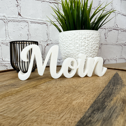 Weißer 3D Schriftzug „Moin“ aus PLA, stehend auf Holzuntergrund vor Ziegelwand mit Pflanze im Hintergrund