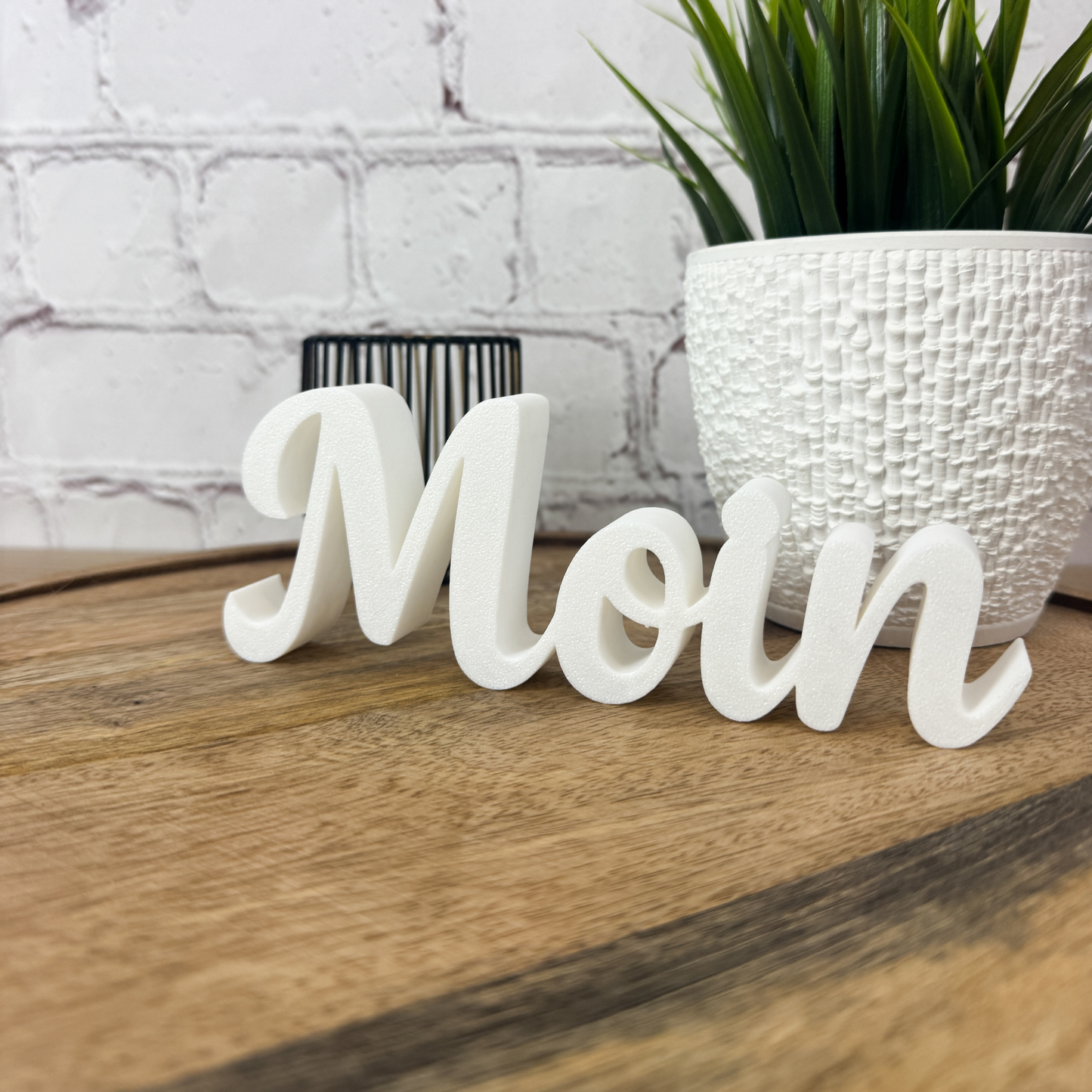 Weißer 3D Schriftzug „Moin“ aus PLA in Seitenperspektive, auf Holzuntergrund neben Pflanzentopf platziert