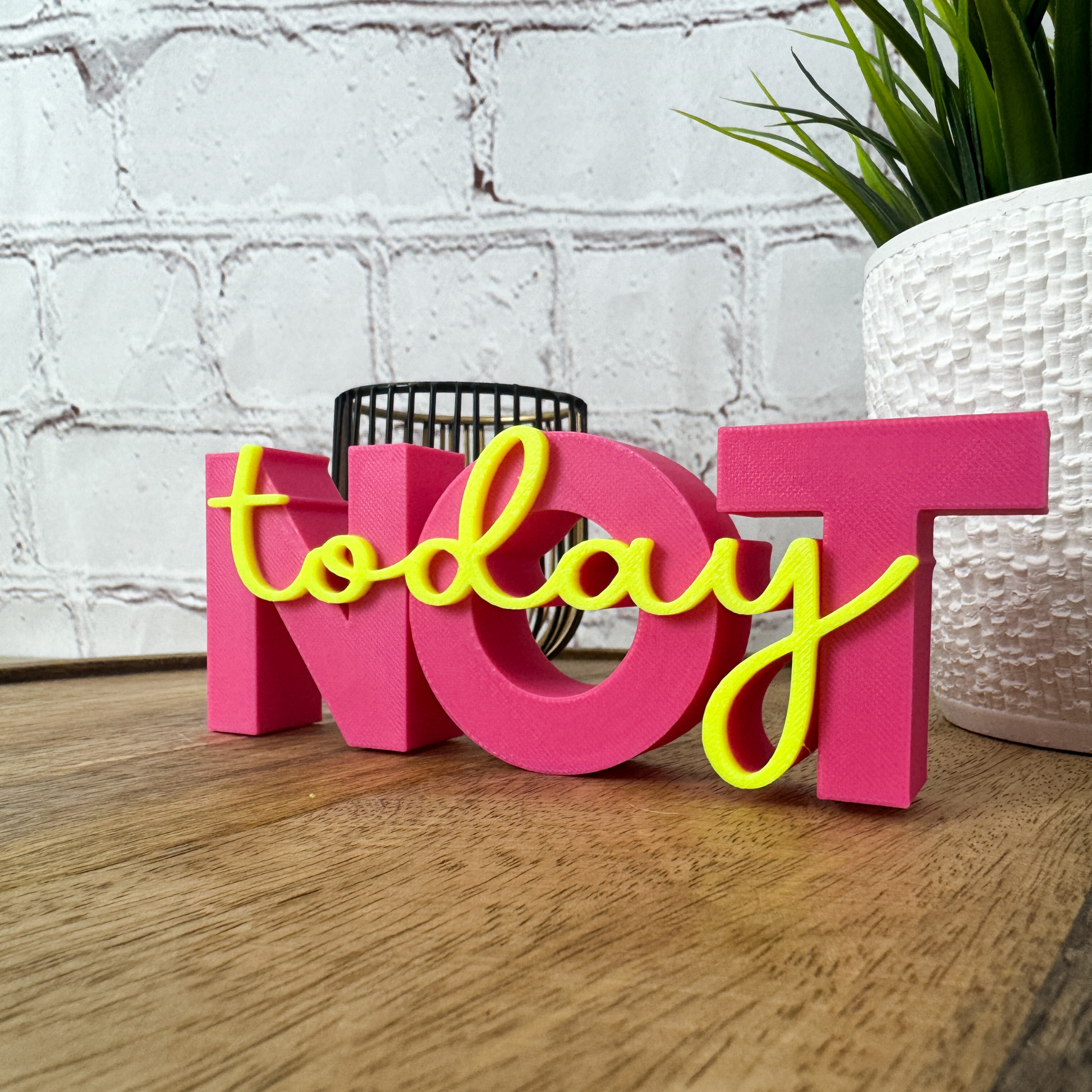 3D Schriftzug „NOT“ in Pink mit gelbem, aufgesetztem Wort „today“, stehend auf Holzuntergrund vor Ziegelwand