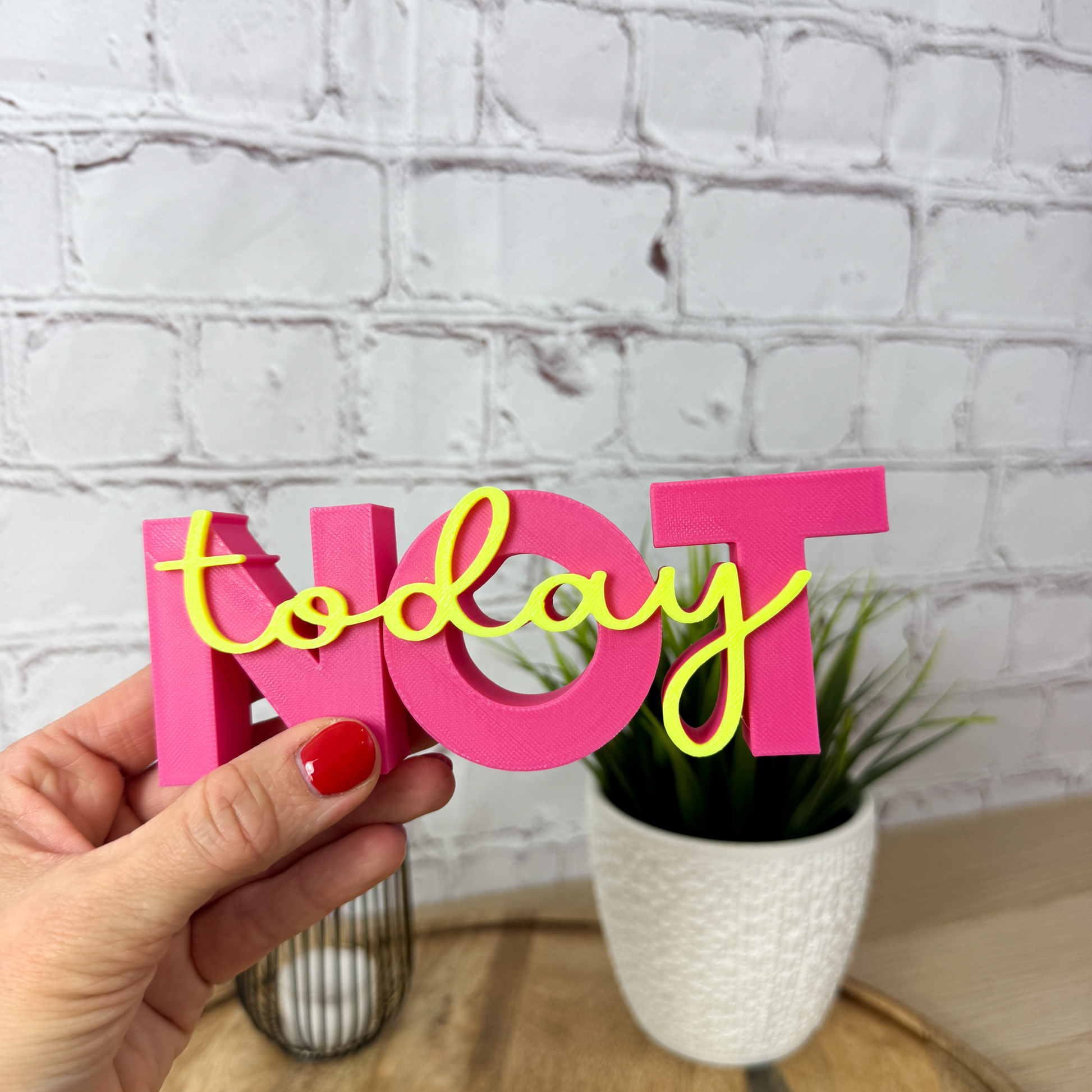 Pinker 3D Schriftzug „NOT“ mit gelbem Wort „today“, frontale Ansicht auf Holzfläche mit Pflanze im Hintergrund