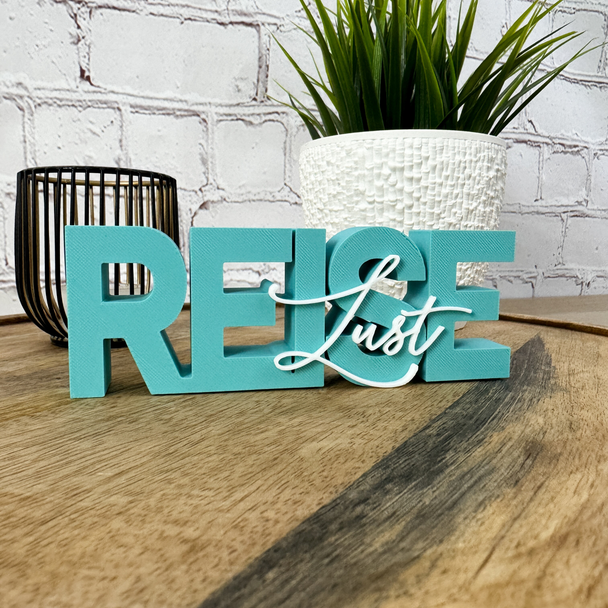 3D Schriftzug „REISE“ in Türkis mit aufgesetztem Wort „Lust“ in Weiß, stehend auf Holzuntergrund vor Ziegelwand