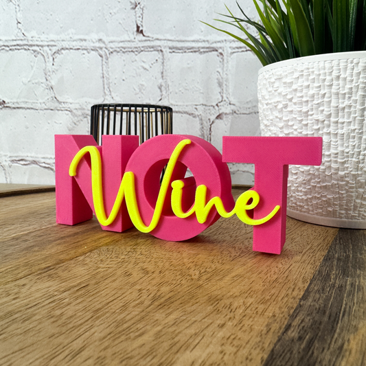 Freistehender 3D Schriftzug „Wine not“ aus PLA in Pink und Neon-Gelb, dekorativ auf Holztisch vor Pflanze platziert