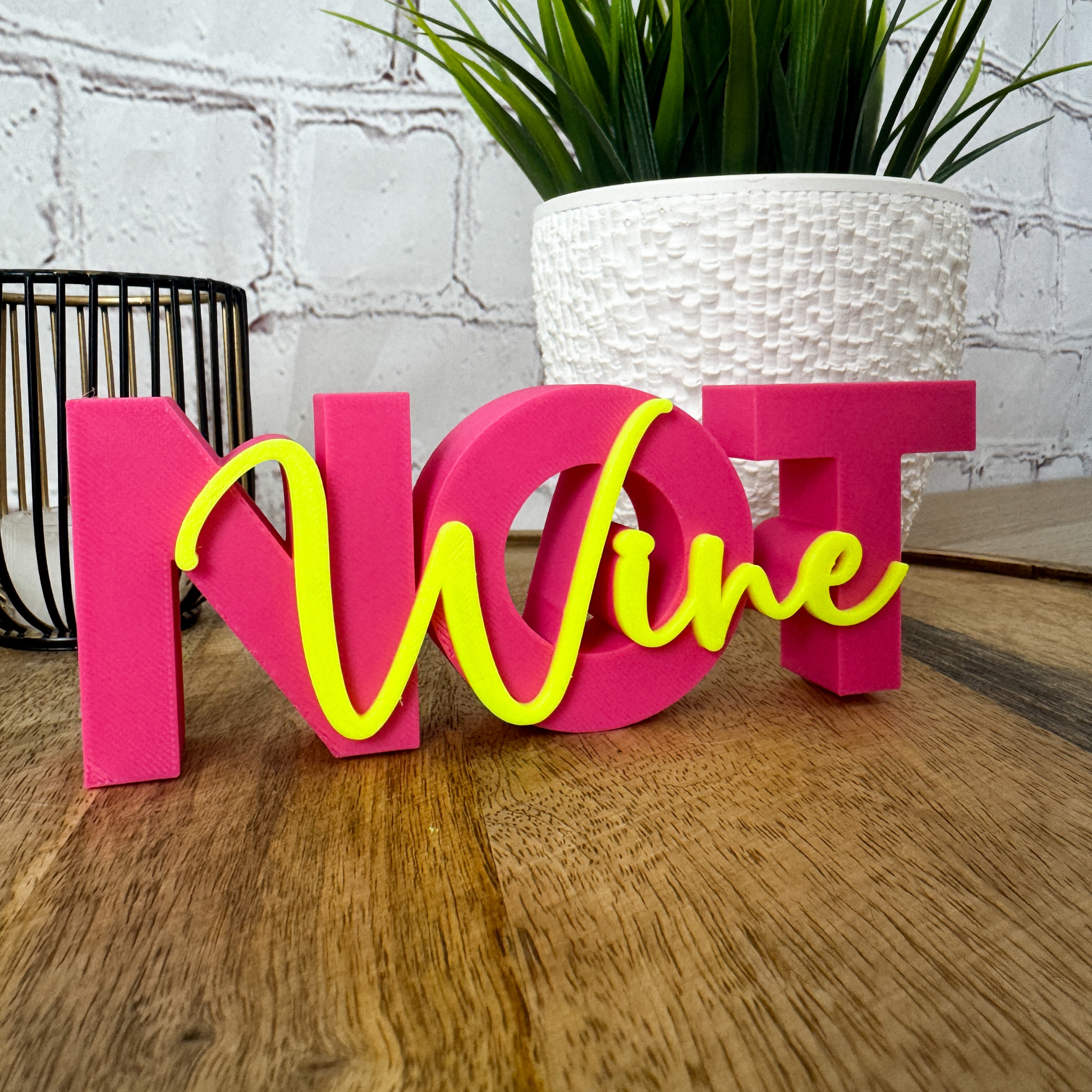 Pink-gelber 3D Schriftzug „Wine not“ aus PLA, als Geschenkidee zum Geburtstag, dekorativ vor Pflanze und Korb arrangiert