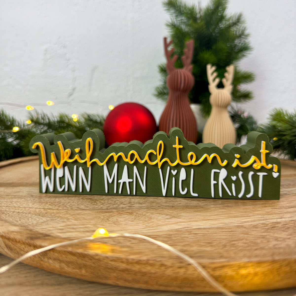 3D Spruch „Weihnachten ist, wenn man viel frisst" als Tischdeko zwischen Christbaumkugel und Rentierfiguren