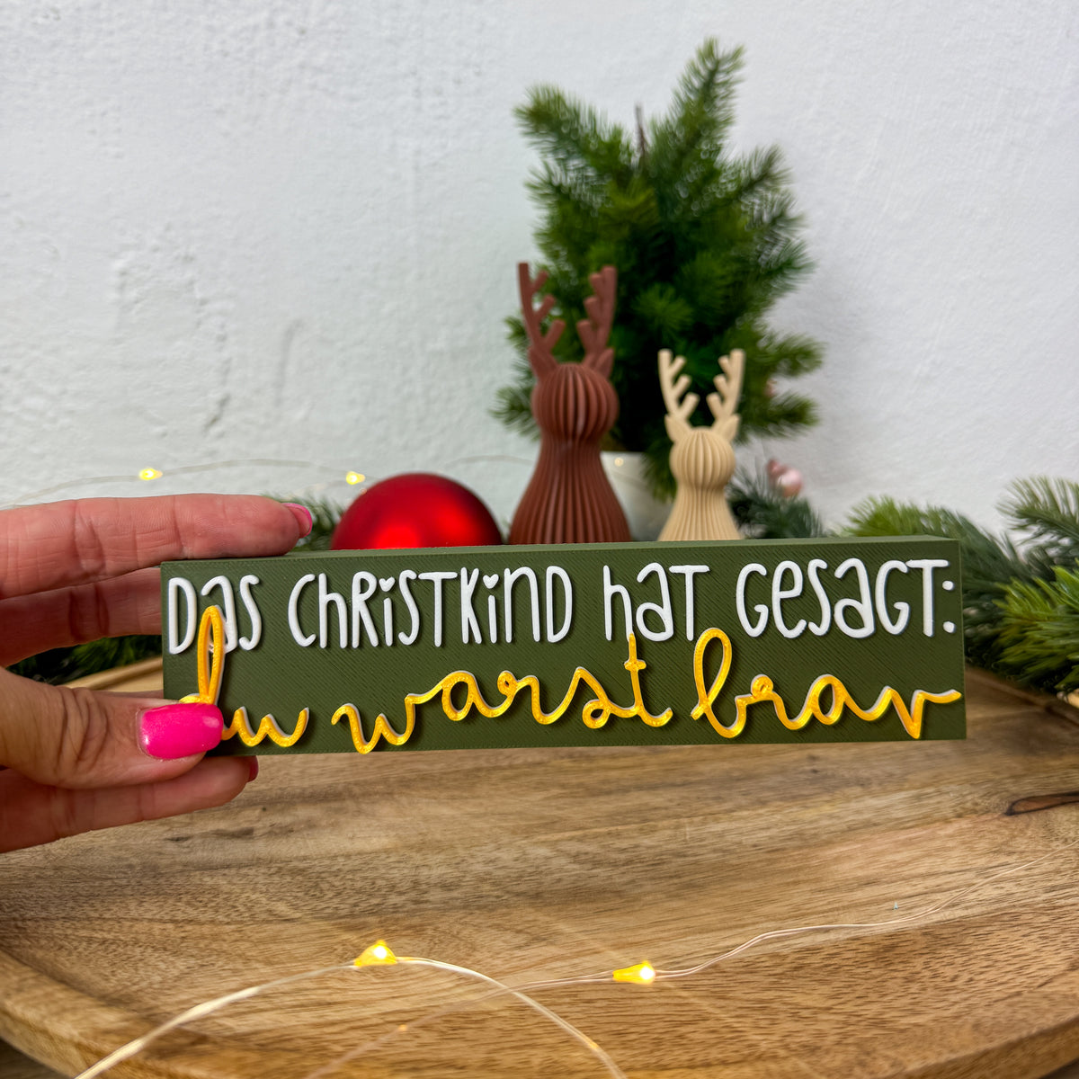Christkind-Spruch-Schild in Weiß und Gold in einer Hand gehalten als Größenvergleich vor Weihnachtsdeko